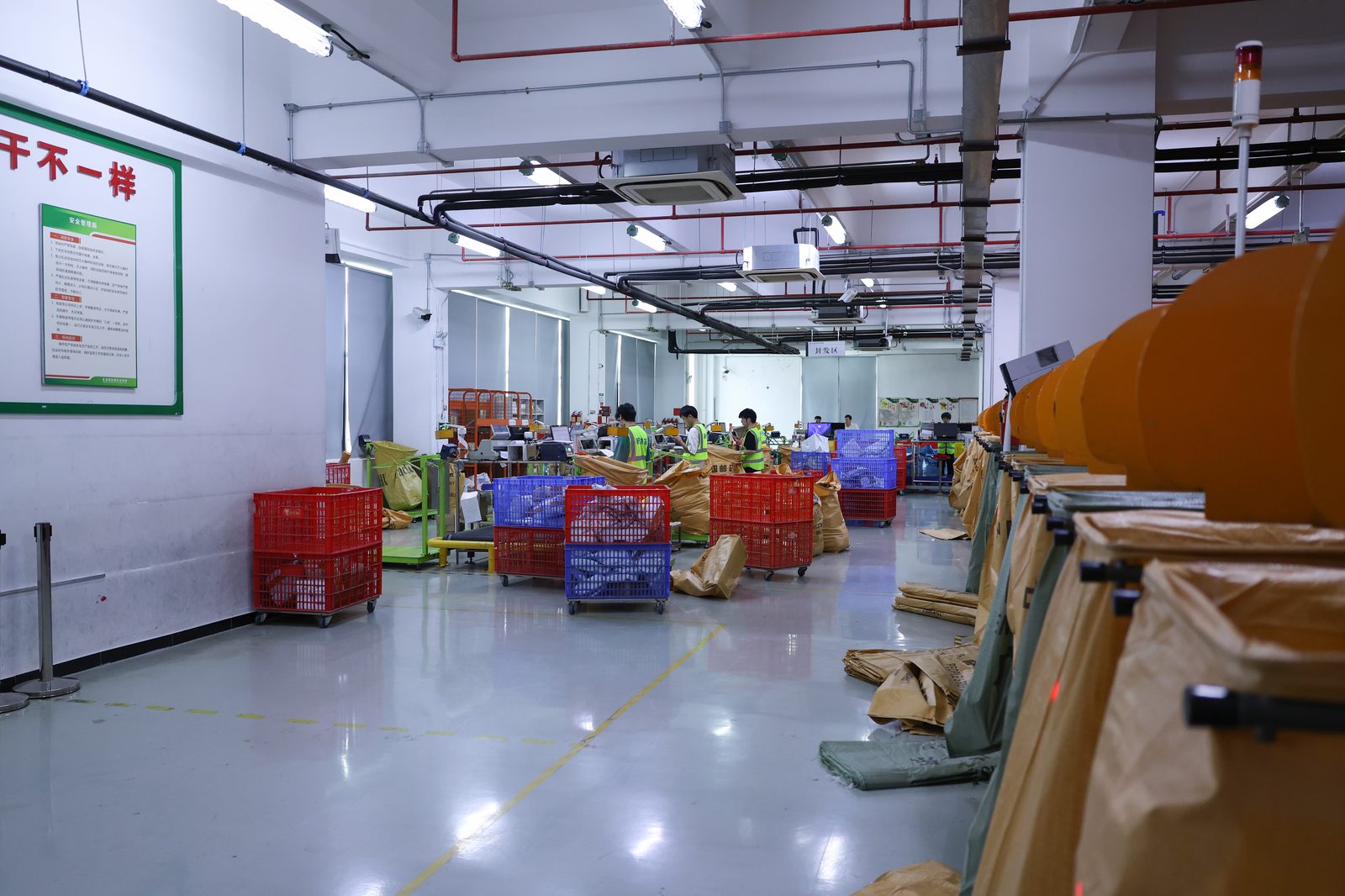 A dispatch area with yellow lane markings and retractable belt barriers. Red and blue plastic crates sit on castors. Four or five workers in hi-vis vests stand at benches with monitors and printers. On the right, open sacks on frames are lined with kraft paper. A sign on the back wall reads dispatch area in Chinese.