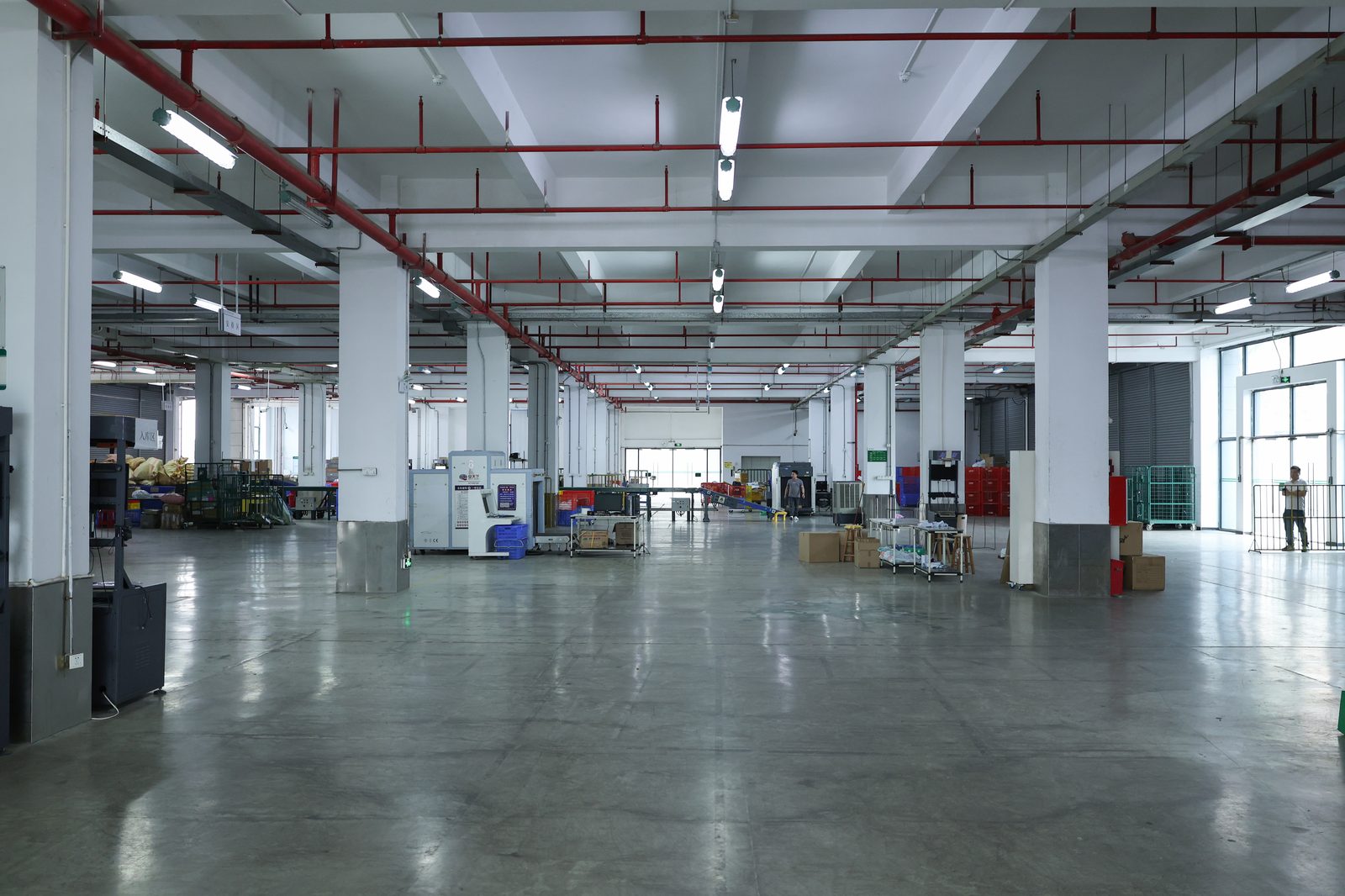 A wide warehouse floor under red sprinkler pipes, with a scanner, wheeled packing carts and green roll cages ranged along the far wall.