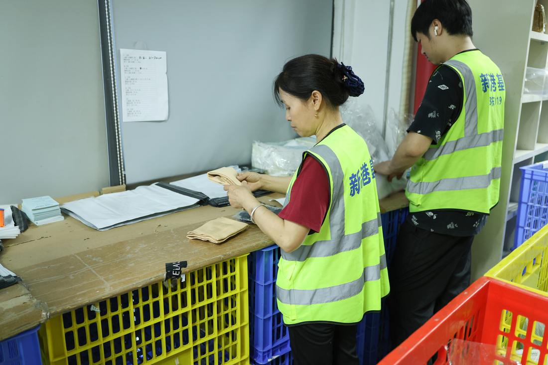 Two workers in hi-vis vests at a wooden bench. The woman in front folds a beige fabric item; the man behind handles clear poly bags. Yellow, blue and red crates are stacked under and beside the bench. White mailers and paperwork lie on the surface, a printed sheet is taped to the wall, and shelving stands to the right.