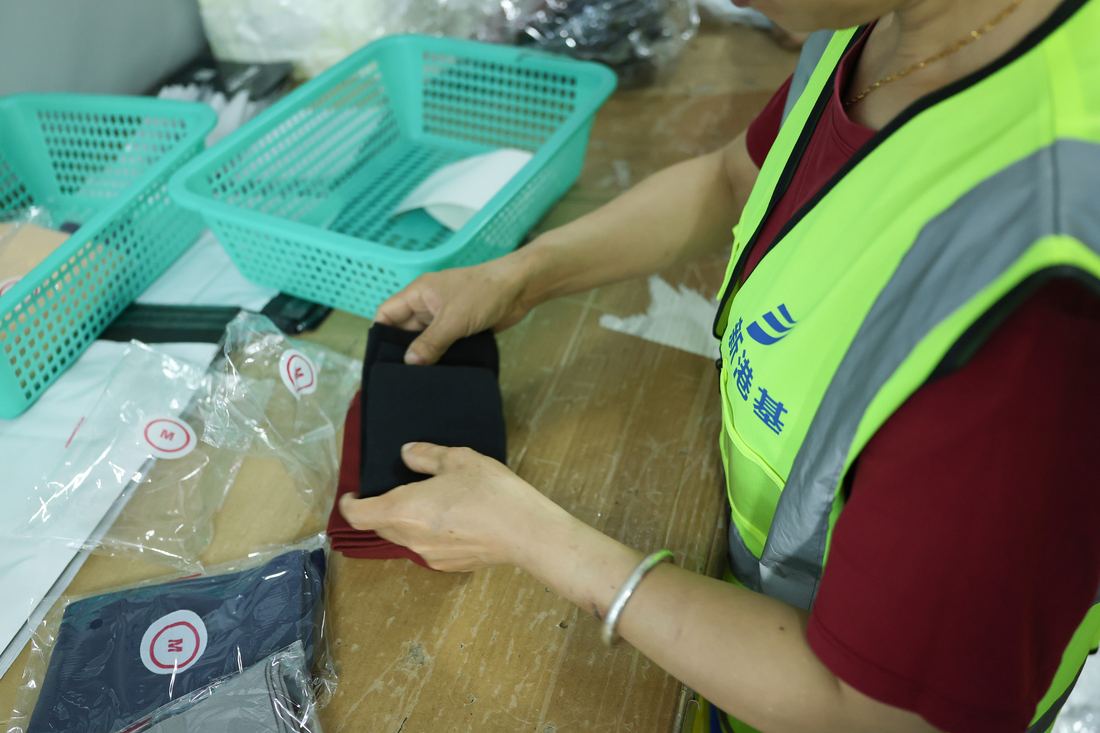A packer folds a dark garment at a wooden bench beside clear poly bags whose round labels show different size markings, with green sorting baskets alongside.