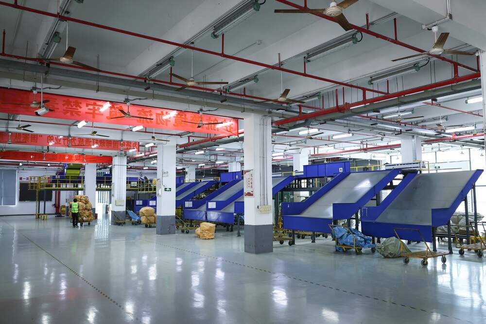 A wide warehouse hall with blue steel chutes in two rows, red overhead banners, ceiling fans, and a single worker in a hi-vis vest pushing a trolley of sacks along the far aisle.