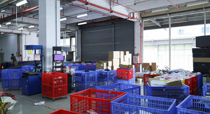 Indoor loading area with a closed roller shutter door: stacked red and blue crates, cardboard cartons piled near the doorway, a scanning station on the left and a pile of packed parcels on the right.