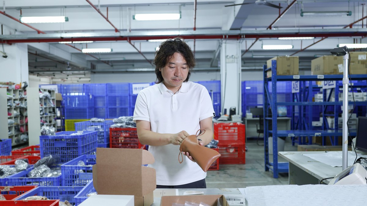 A worker in a white polo shirt turns a tan leather pouch over in his hands at a packing bench, with stacked blue and red crates and boxed stock on shelving behind him.
