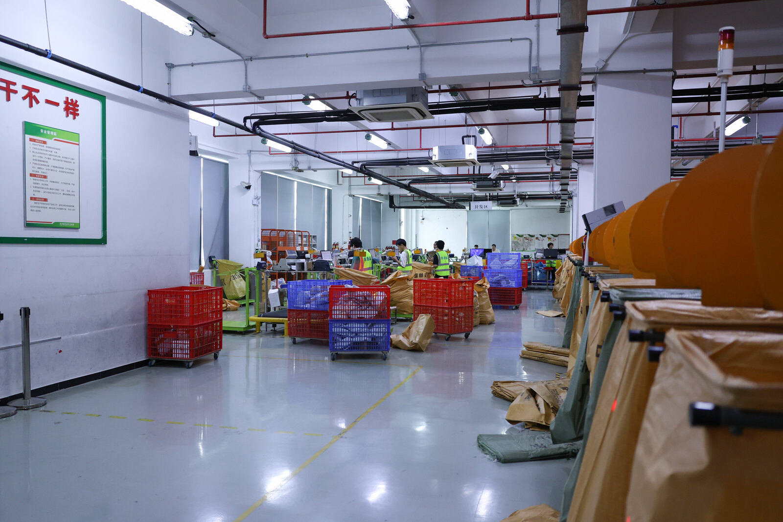 Warehouse staff work among wheeled sorting cages, parcel sacks and orange dispatch chutes on a clean fulfilment floor.