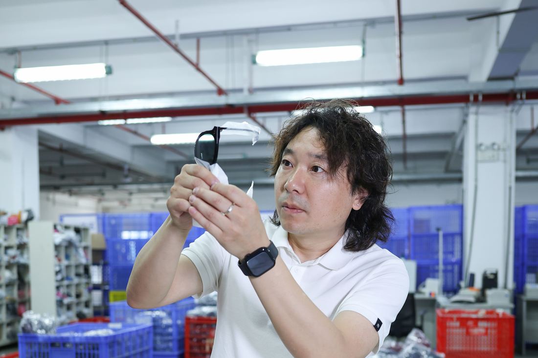 A worker holds a pair of sunglasses up toward the ceiling lights and looks through the lenses, standing over blue and red crates of stock.