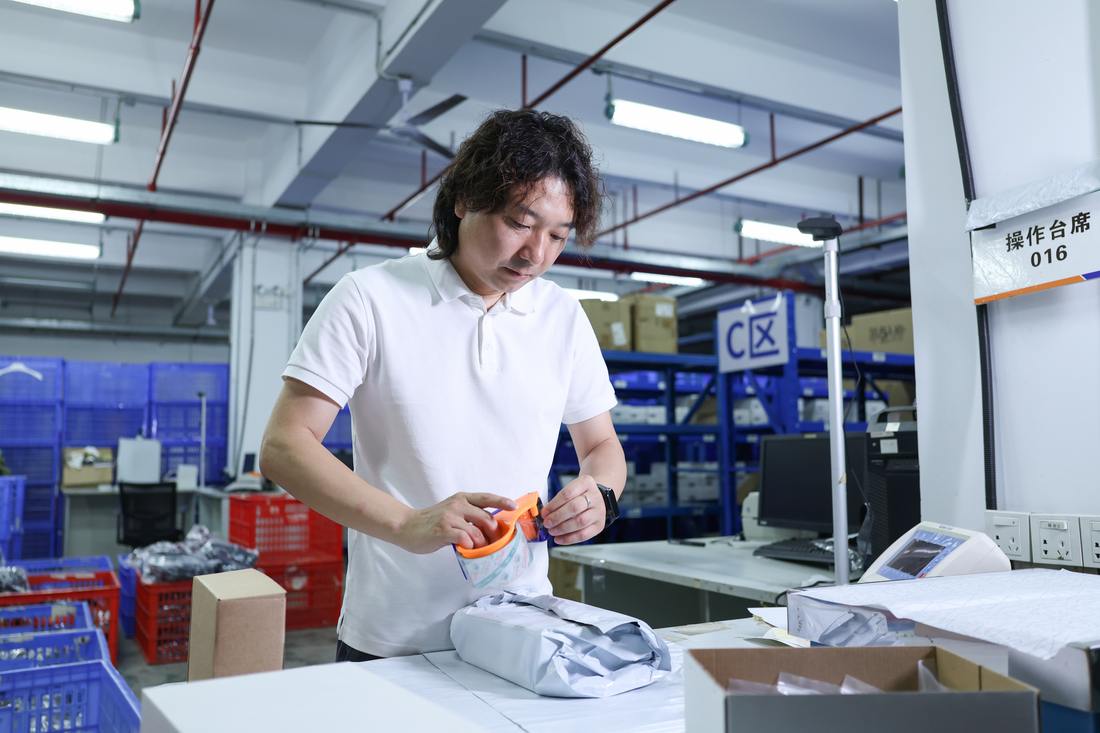 A worker cuts open a small poly-bagged item with an orange safety cutter at workstation 016, an opened grey mailer lying on the bench in front of him and blue shelving behind.