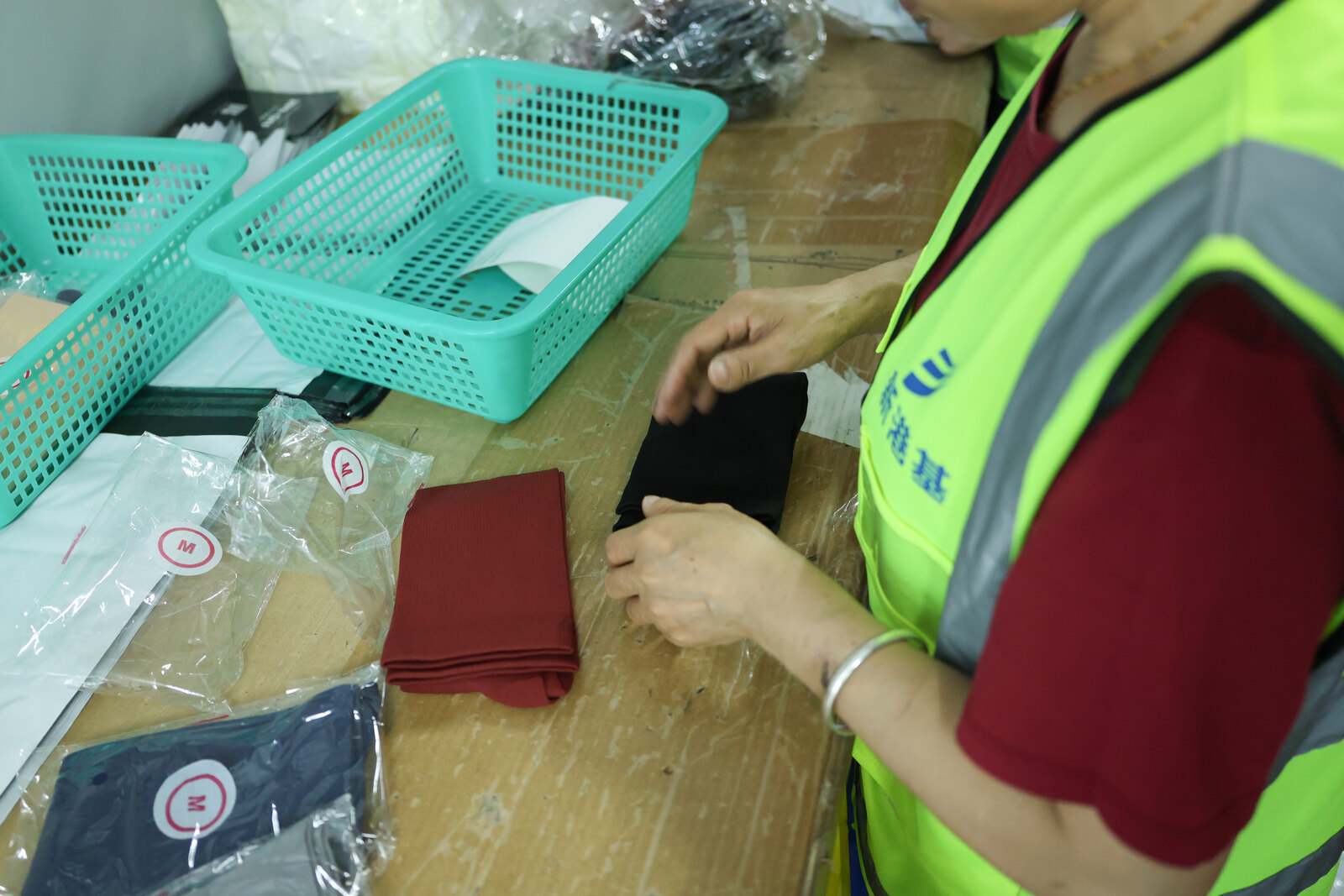 A warehouse worker folds and sorts small fabric items beside labelled poly bags and green picking baskets at a packing bench.