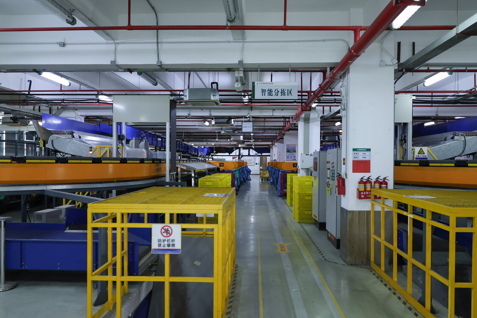 A clear aisle between automated orange and blue parcel sorting conveyors, with yellow safety barriers and collection cages.