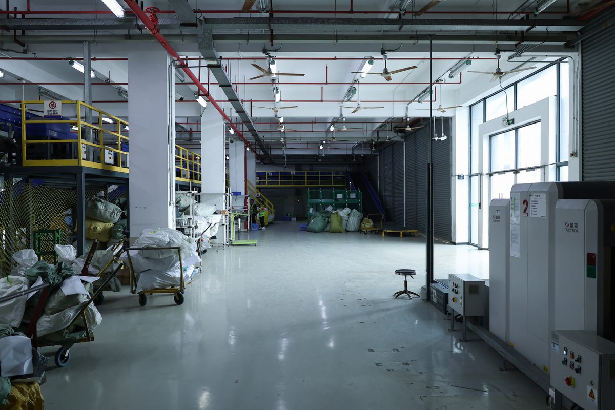 A warehouse interior with a yellow mezzanine rail and stairs on the left and large industrial ceiling fans overhead alongside red sprinkler pipes. Woven bulk bags in green and white are stacked on trolleys along the left wall. On the right stands a NUCTECH X-ray scanner marked with the number 2. A roller shutter and tall windows let in daylight; green mesh cages sit in the middle distance with one worker in hi-vis further back.