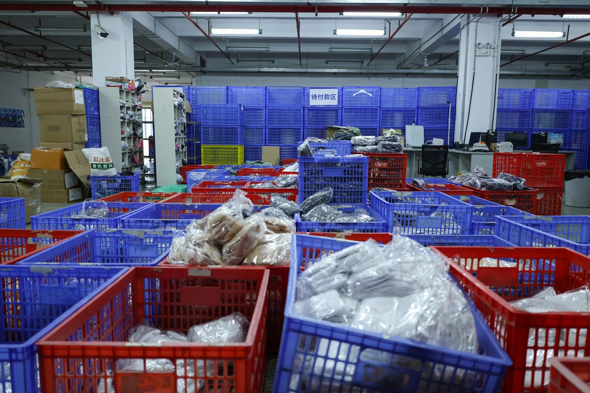 Rows of red and blue plastic crates filled with poly-bagged goods, stacked several deep across the floor. A sign on the wall behind reads pending payment area in Chinese. Shelving on the left holds more bagged items, cartons are stacked beside it, and a desk with monitors sits at the back right.
