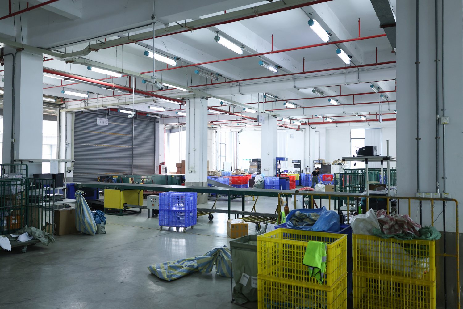 A warehouse floor with a roller shutter door on the left and a long conveyor running across the frame. Blue and yellow plastic crates stand in the foreground, woven sacks beside them, and one worker is visible in the distance. Red sprinkler pipes and fluorescent strips run overhead.