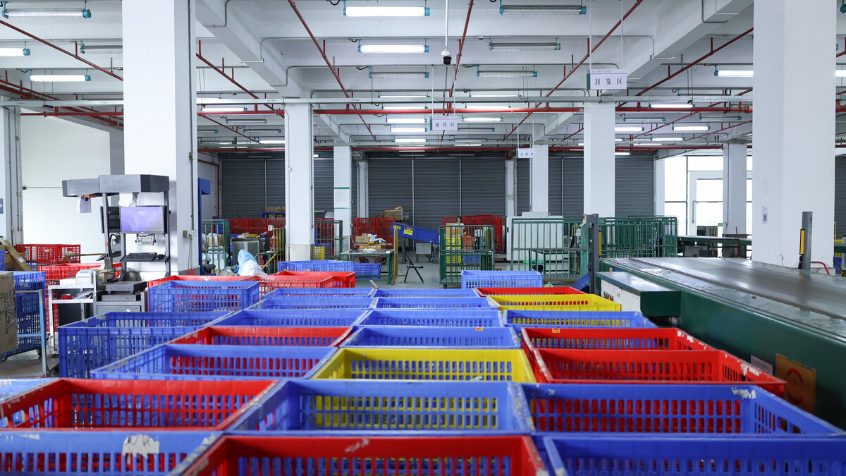 Rows of red, blue, and yellow plastic sorting crates stand beside a conveyor inside a warehouse.