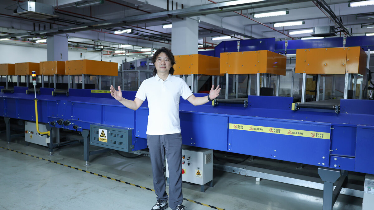A man in a white polo shirt stands beside a long blue and orange conveyor and sorting machine inside an ASG warehouse.