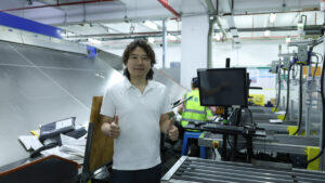 A man in a white polo shirt stands at an ASG warehouse workstation beside a monitor, conveyor rollers, stacked packaging materials, and a worker in a high-visibility vest.