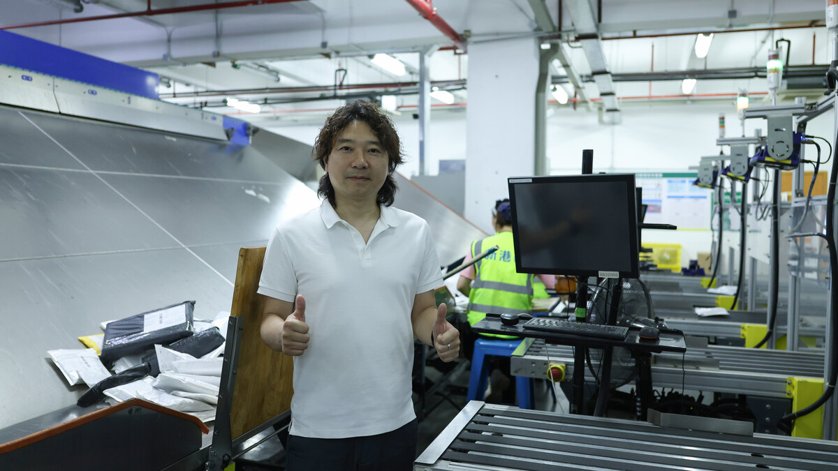 A man in a white polo shirt stands at an ASG warehouse workstation beside a monitor, conveyor rollers, stacked packaging materials, and a worker in a high-visibility vest.