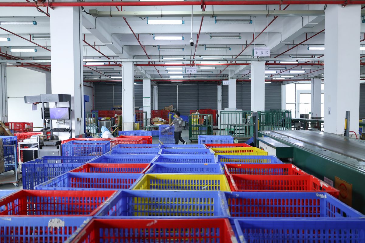 Empty red, blue and yellow plastic crates arranged in rows in the foreground. A worker walks through the middle distance carrying something. A green conveyor runs along the right. Roller shutter doors are closed at the back, with hanging signs above marked dispatch area in Chinese.