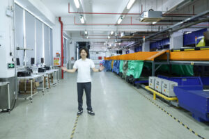 ASG staff member standing in a bright warehouse aisle beside blue and orange automated sorting conveyors, with packing equipment extending into the background.