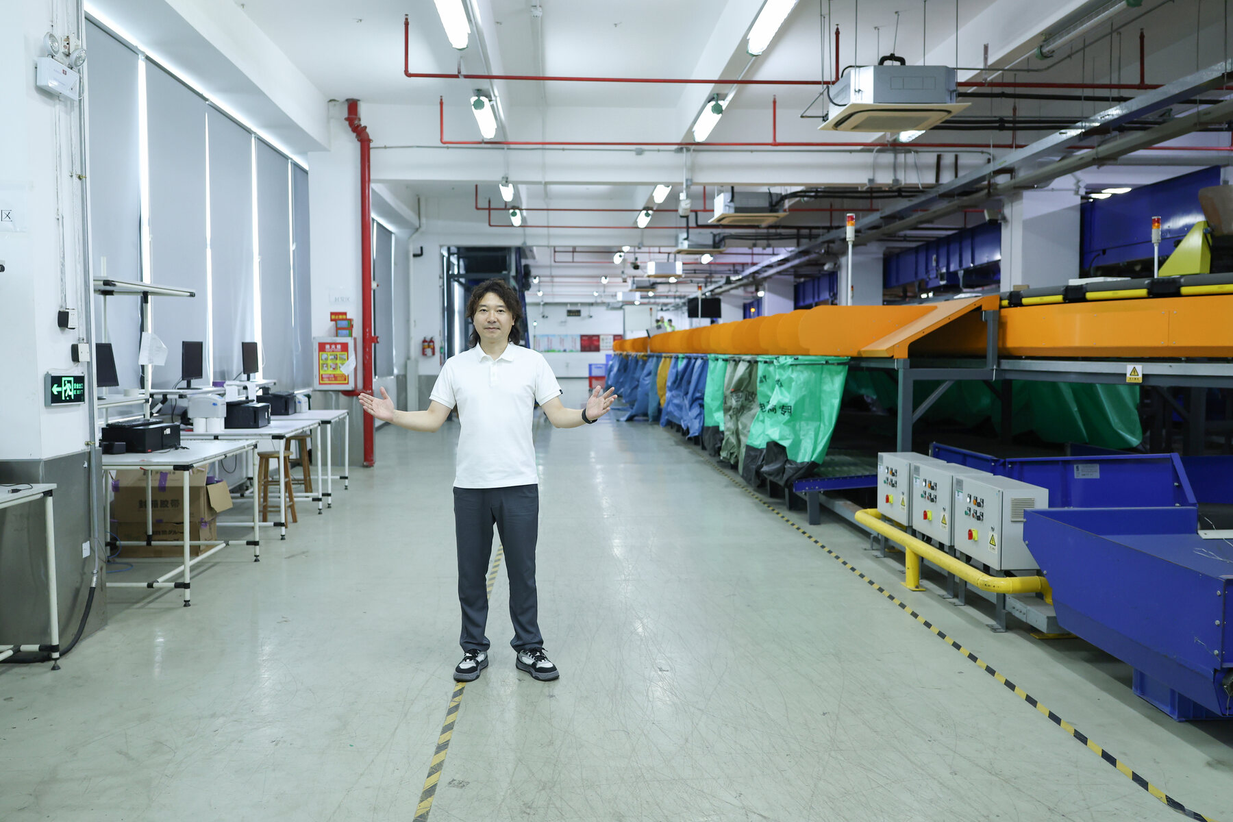 ASG staff member standing in a bright warehouse aisle beside blue and orange automated sorting conveyors, with packing equipment extending into the background.