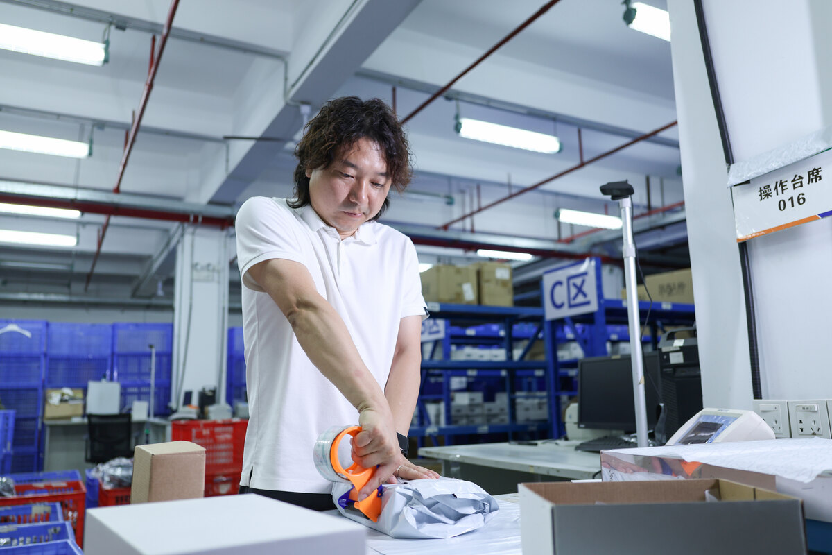 A worker closes a parcel at a warehouse packing bench beside cartons and sorting bins.
