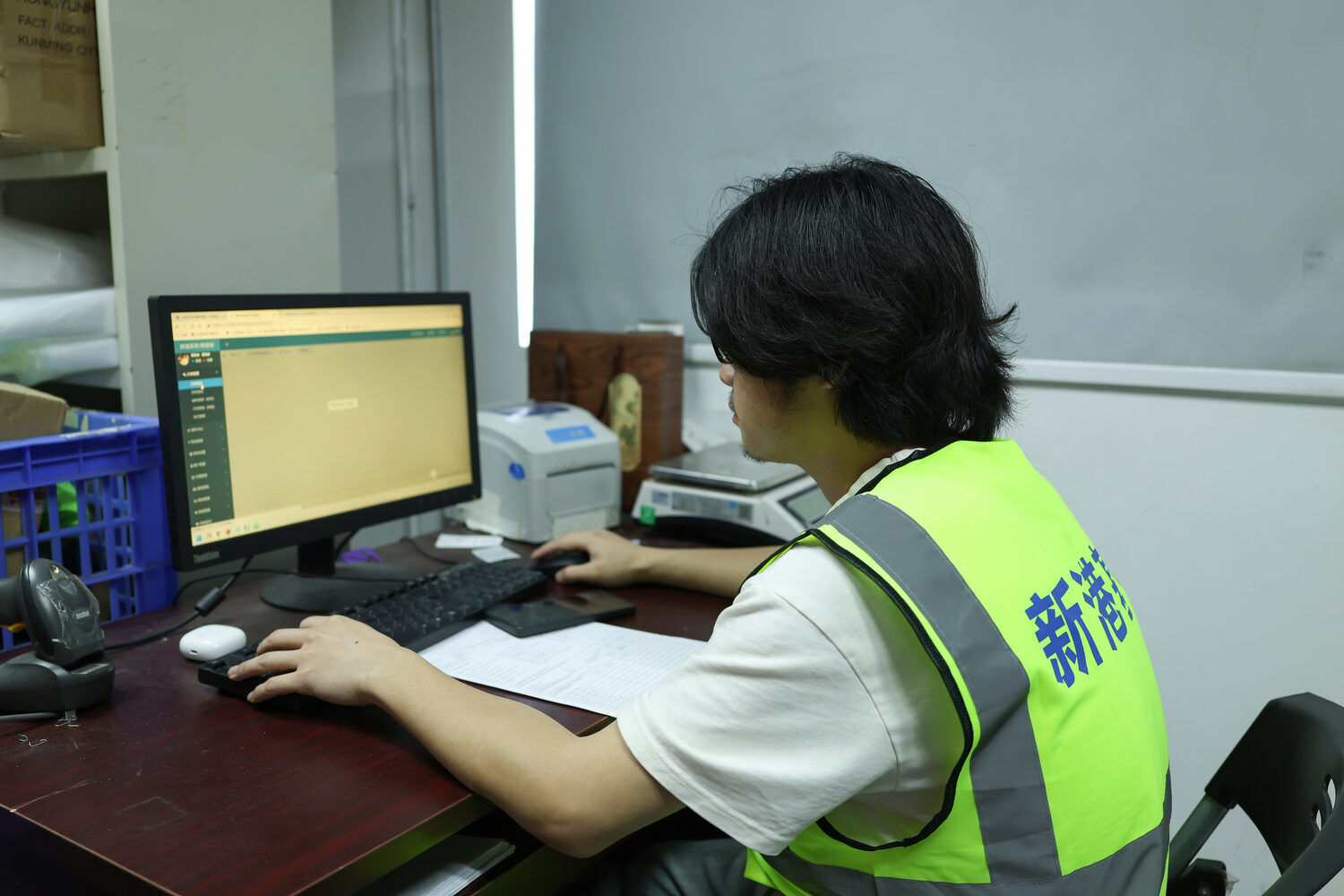 ASG warehouse worker seated at a computer workstation with a barcode scanner, label printer and paper order sheet.