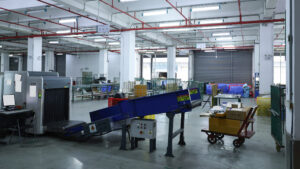 A security-screening machine with a blue conveyor beside a trolley loaded with parcels.