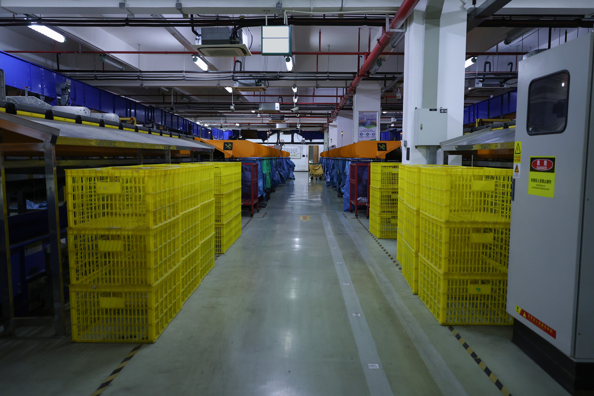 Stacks of yellow crates line a clear aisle beneath sorting chutes, with bag stands farther back.