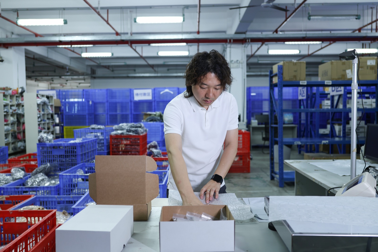 A man placing an item in a cardboard box at a warehouse packing bench with plastic baskets behind him.