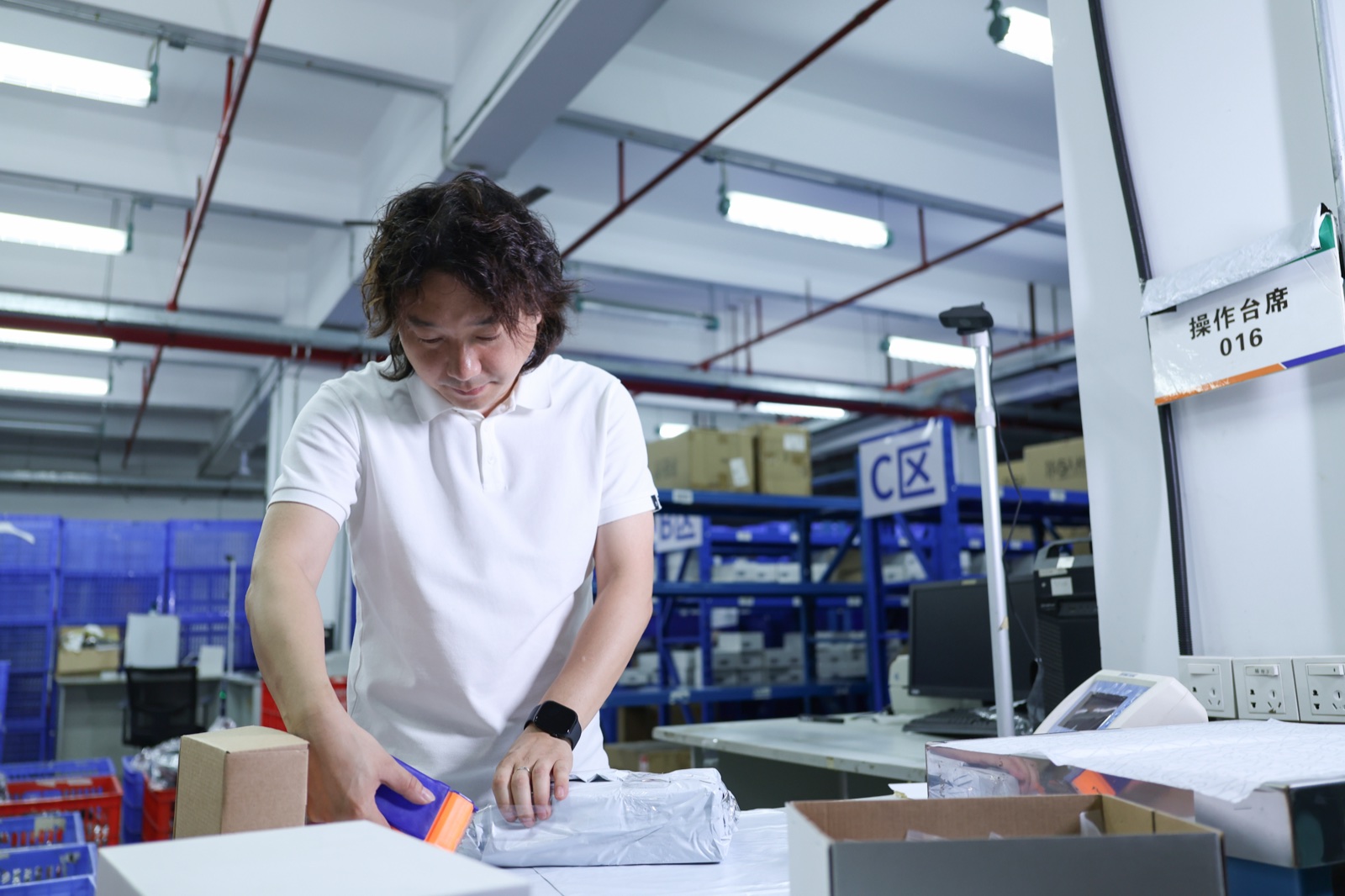 A man handling a wrapped parcel at a warehouse workbench beside cartons and racks.
