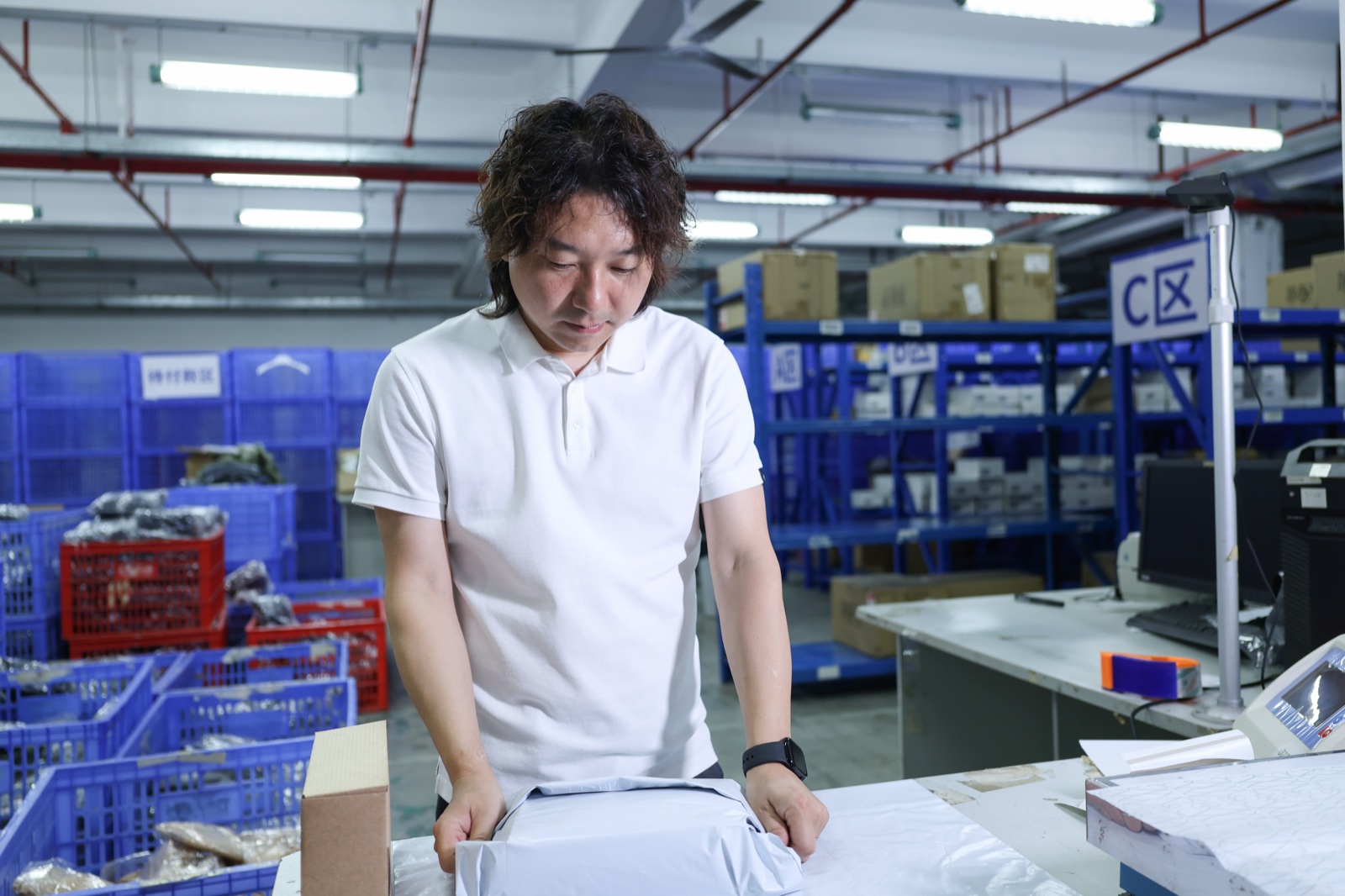 A man at a warehouse packing bench with cartons on blue storage racks behind him.