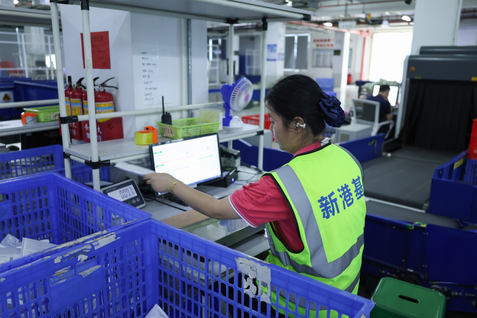 A warehouse worker beside a computer and scale with blue plastic baskets nearby.