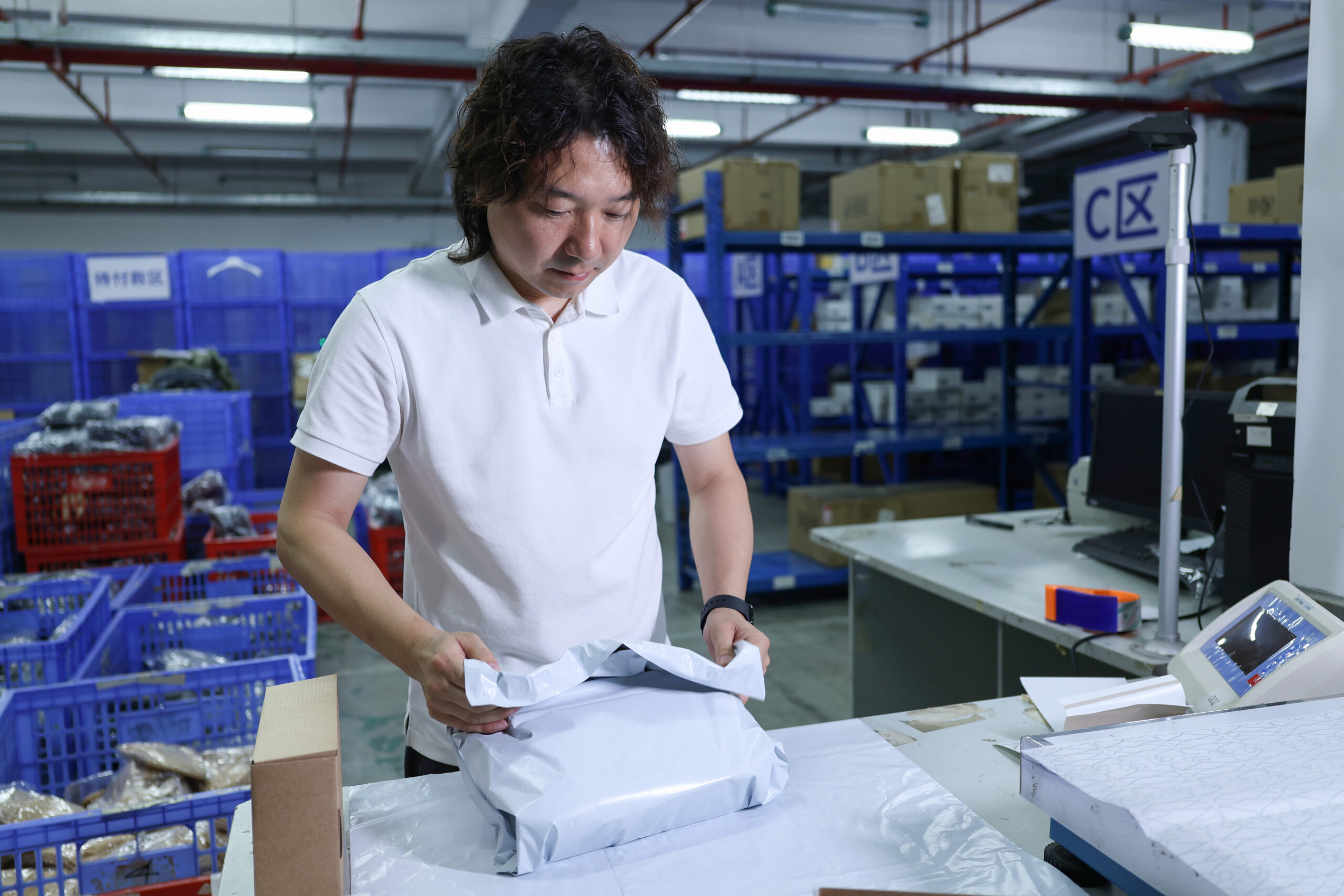A person placing a packaged item into a white mailing bag at a warehouse bench.