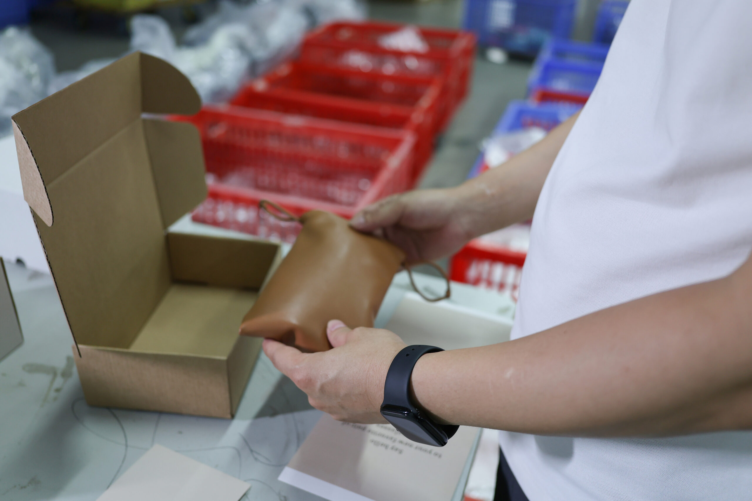 Hands holding a tan pouch beside an open cardboard box at a packing station.