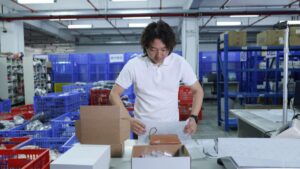 Person wrapping a small brown pouch in white packing paper at a warehouse packing table, with open boxes and plastic crates nearby; featured image for this guide to Printful and your own stock in one Shopify order