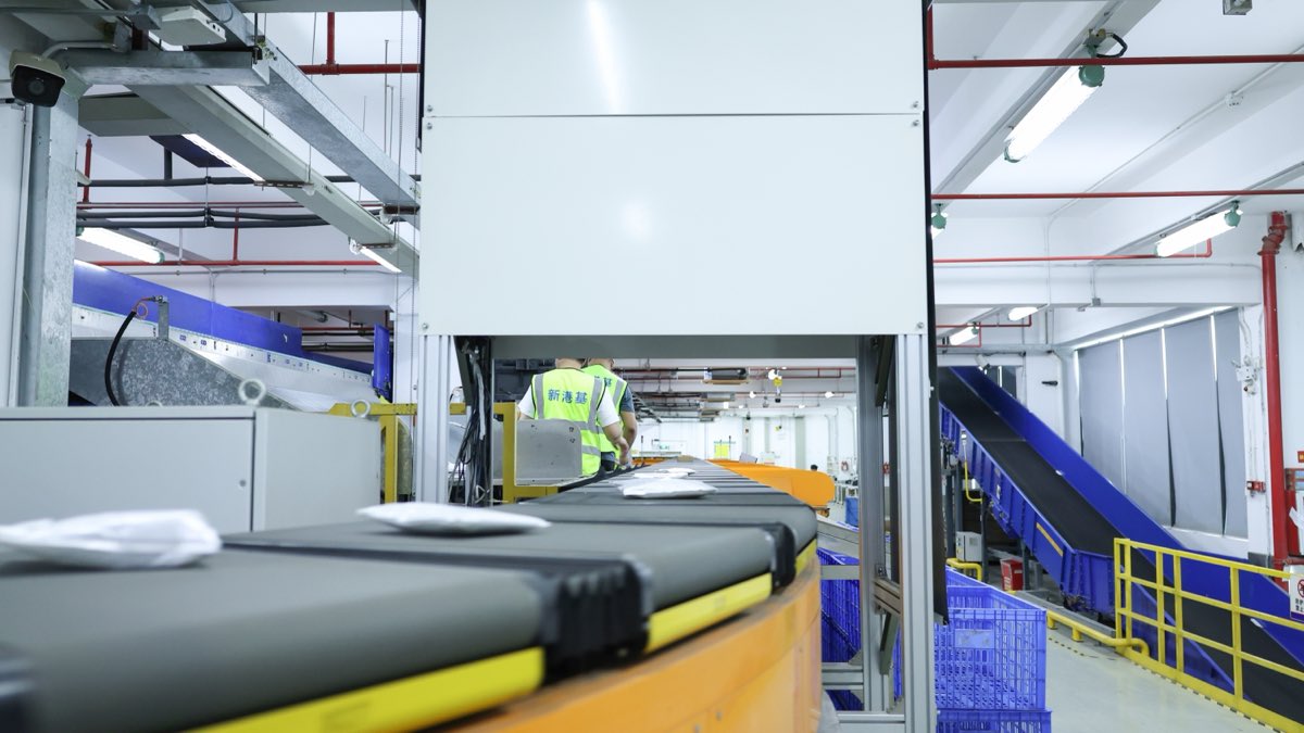 Flat sealed parcels on the grey trays of a sorting conveyor, with blue crates, an inclined conveyor and two workers in hi-vis vests in the background.