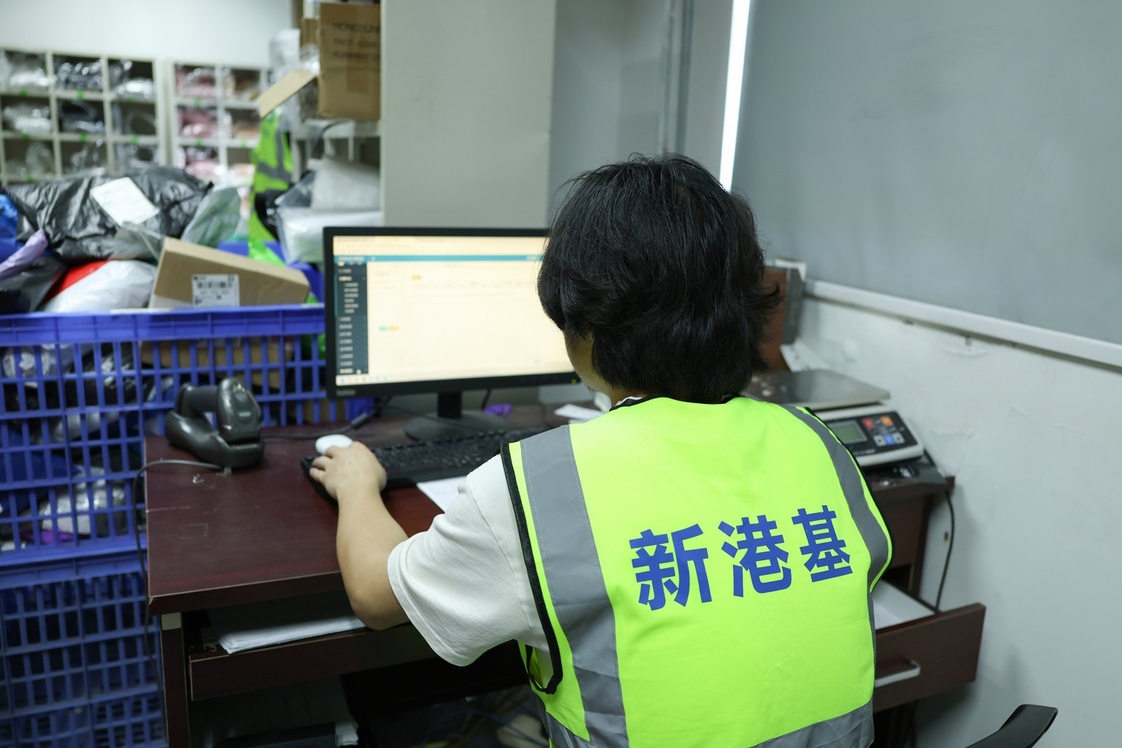 A worker in a bright vest sits at a warehouse computer beside a scanner, scale and bins of parcels.