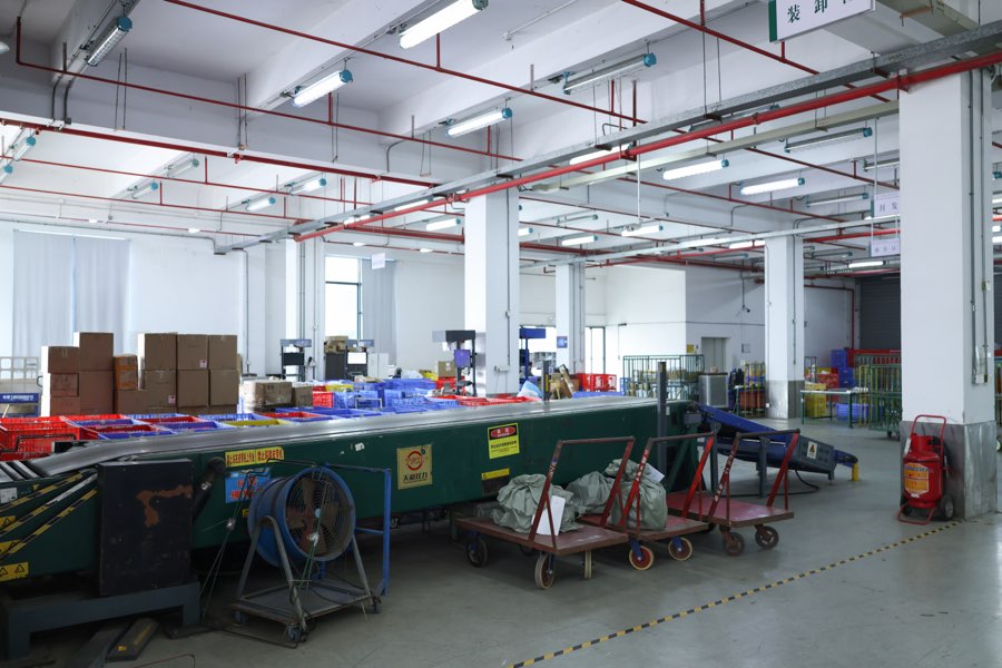 A loading area with a long green conveyor, stacked cartons, red and blue crates, and carts holding filled sacks under ceiling lights.