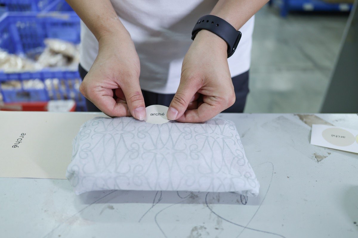 Hands pressing a round printed sticker onto a tissue-wrapped item on a packing table, with printed paper pieces beside it.