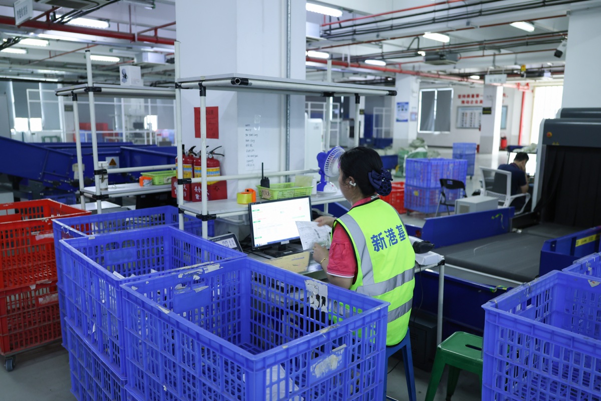 A worker in a bright vest sits at a computer workstation among large blue plastic totes.