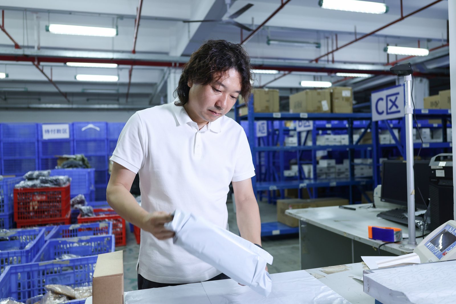 Janson inspecting a white packaged item beside an outbound warehouse workstation and parcel bins.
