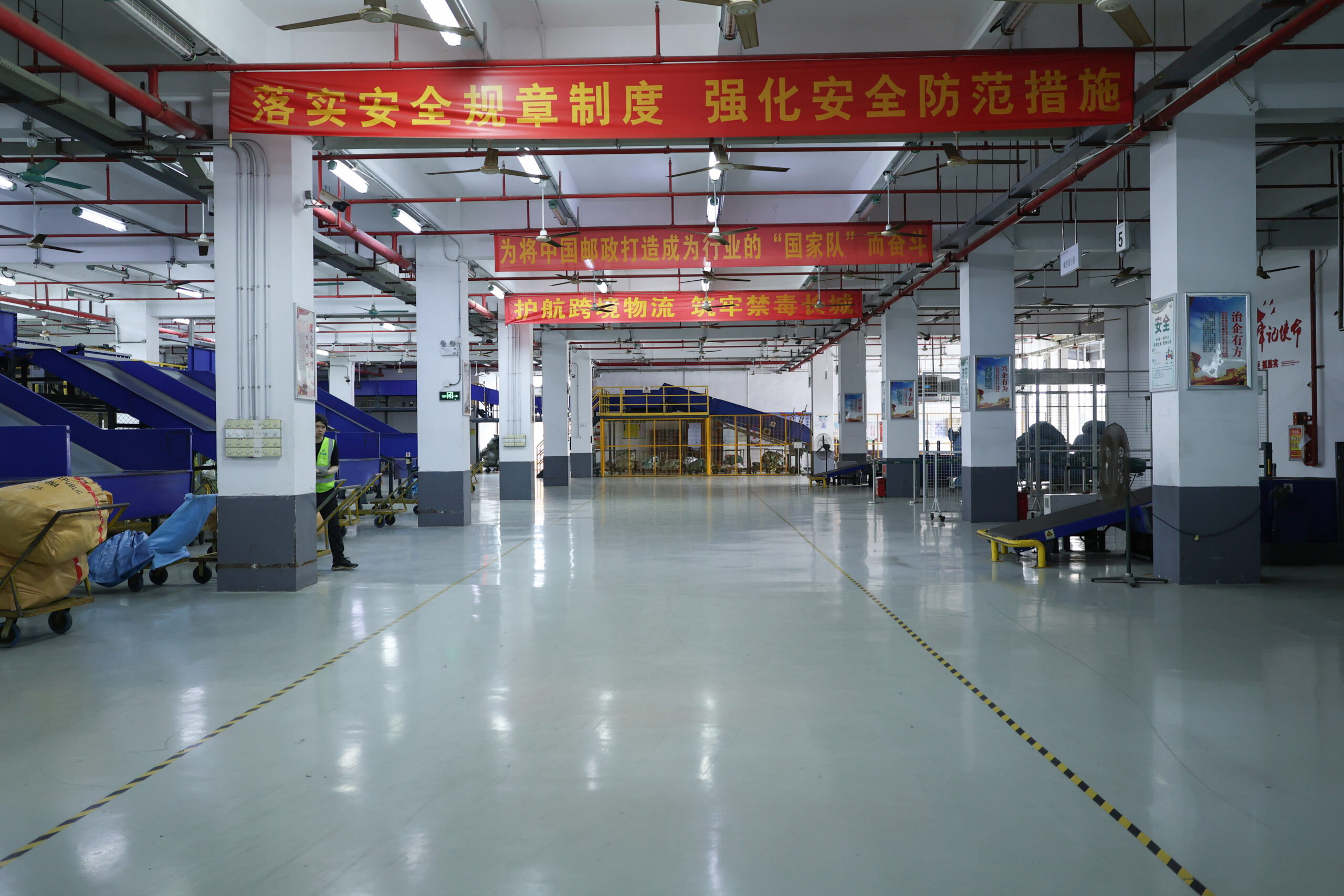 Parcel sorting floor with conveyors, wheeled bags and overhead safety banners in China.